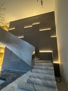 Modern luxury villa staircase with marble steps, textured metal railing, and geometric backlit wall panels in a high-ceiling lobby.