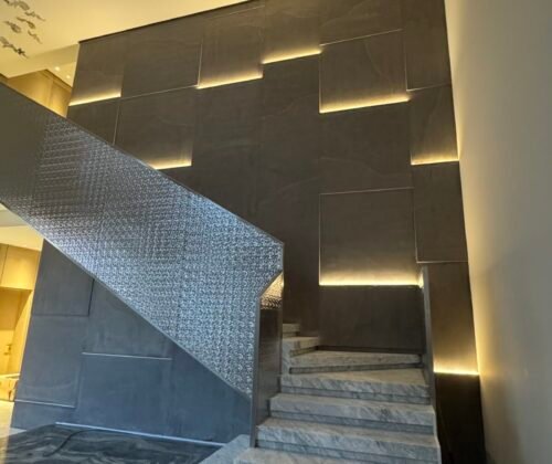 Modern luxury villa staircase with marble steps, textured metal railing, and geometric backlit wall panels in a high-ceiling lobby.